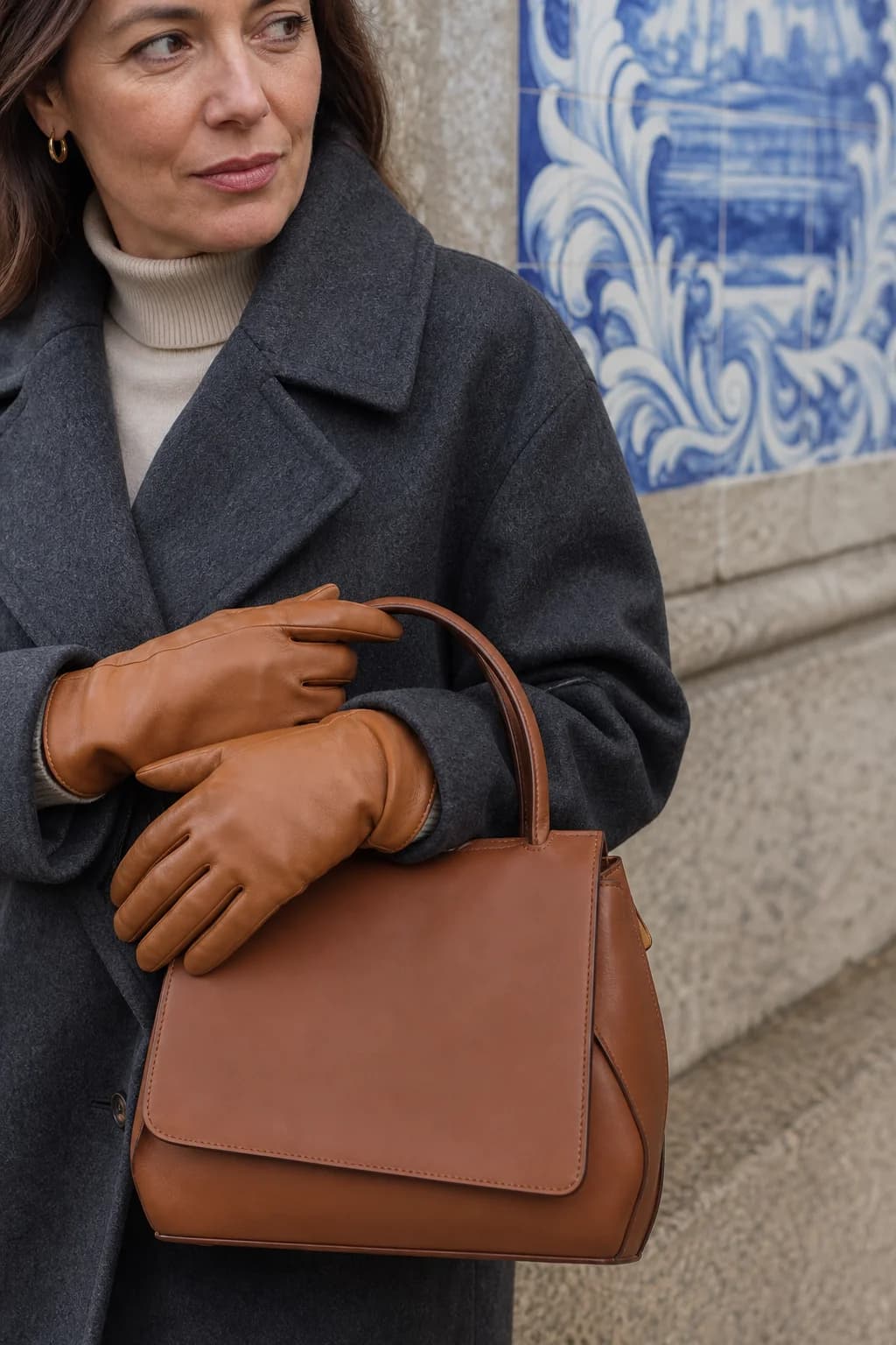 Mãos com luvas de pele a segurar uma mala castanha junto a azulejos
